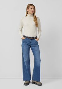 Crème coltrui gecombineerd met blauwe brede jeans en een bruine riem. De outfit heeft een soepele textuur en een relaxte pasvorm.