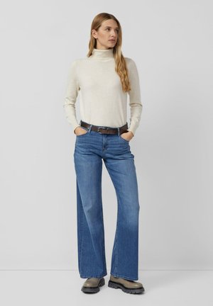 Crème coltrui gecombineerd met blauwe brede jeans en een bruine riem. De outfit heeft een soepele textuur en een relaxte pasvorm.