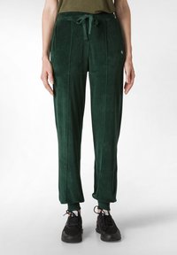 Grüne Velours-Sweatpants mit gebundenem Bund, zwei seitlichen Nähten und taillierten Bündchen. Weicher Stoff, lässiges Design, kombiniert mit schwarzen Sportschuhen.