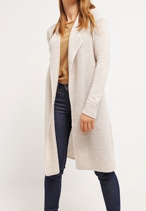 Mujer con un cárdigan largo de punto beige sobre una camisa color canela y vaqueros pitillo azul oscuro, de pie contra un fondo blanco liso.