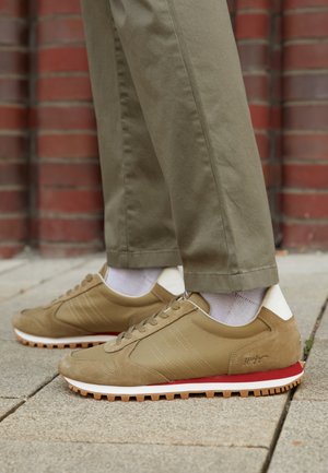Personne portant des baskets beiges avec des chaussettes blanches et un pantalon kaki, debout sur un sol pavé près d'un mur en briques rouges.