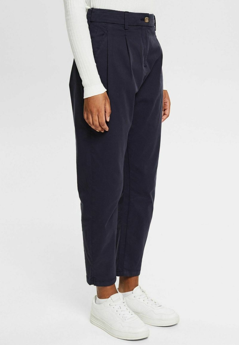 Personne portant un pantalon bleu marine taille haute fuselé avec des plis devant, associé à un haut blanc côtelé à manches longues et des baskets blanches, debout de profil.