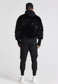 Muž stojící zády, oblečený v černé bundě z umělé kožešiny se nápisem "SIKSILK" na zádech, černých cargo kalhotách, bílých teniskách a černé čepici.