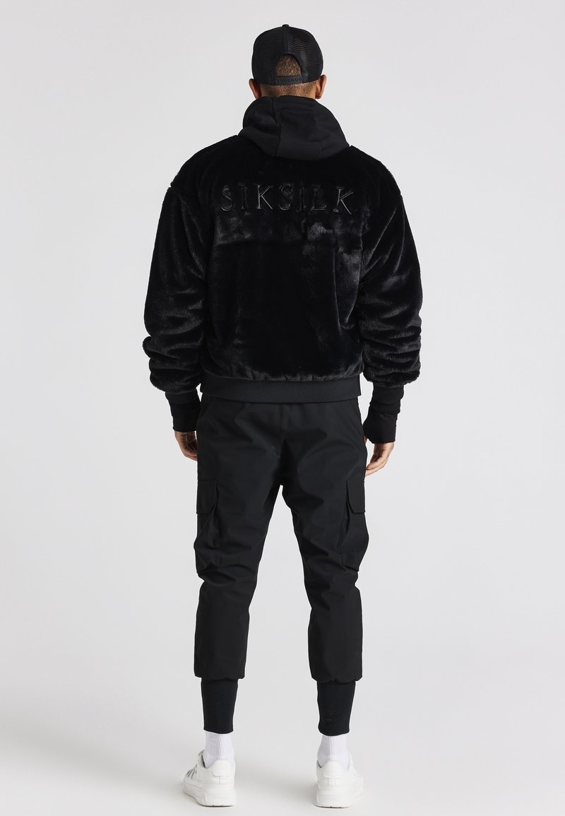 Muž stojící zády, oblečený v černé bundě z umělé kožešiny se nápisem "SIKSILK" na zádech, černých cargo kalhotách, bílých teniskách a černé čepici.