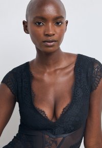 Black lace bodysuit with a deep V-neck, featuring delicate floral patterns and sheer accents. Smooth texture with a fitted design.