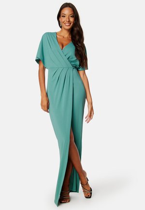 Turquoise maxi jurk, zachte stof, korte mouwen, wikkelstijl lijfje, geplooide details, zijslit, gecombineerd met zwarte strapsandalen.