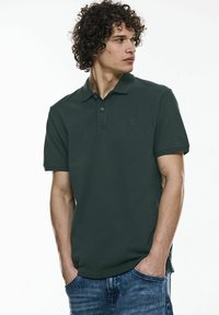 Polo shirt vert foncé en coton, avec une patte de boutonnage à trois boutons et un petit logo sur la poitrine gauche, associé à un jean bleu.