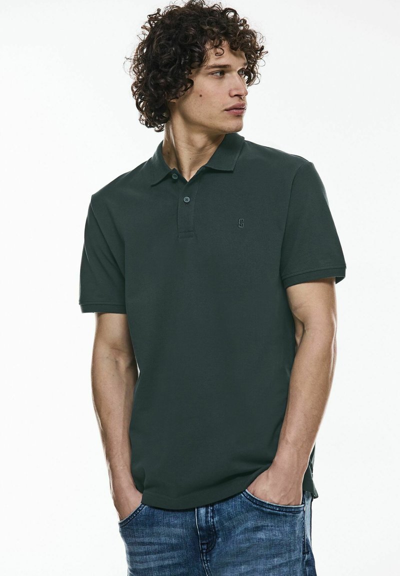 Polo shirt vert foncé en coton, avec une patte de boutonnage à trois boutons et un petit logo sur la poitrine gauche, associé à un jean bleu.