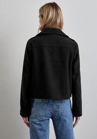 Svart kort jacka med slagkrage och långa ärmar, tillverkad av ett glatt tyg. Buren över ljusblå denimjeans, visande bakvy.