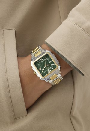 Pols met een tweekleurig zilver- en gouden metalen horloge met een groen vierkant wijzerplaat, Romeinse cijfers en een datumaanduiding, tegen een beige stof.