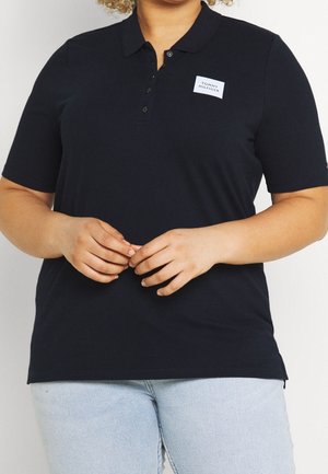 Personne portant un polo bleu marine avec le logo "Tommy Hilfiger" sur la poitrine et un jean bleu clair, les mains jointes à la taille.