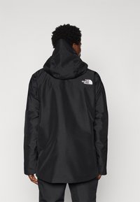 Schwarze wasserdichte Jacke mit Kapuze, entworfen mit einem lockeren Schnitt, versehen mit einem weißen Logo auf dem oberen Rücken und glatter Textur.