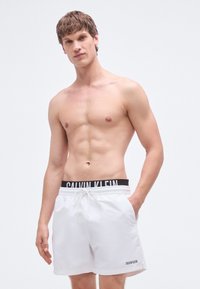 Λευκά μαγιό σορτς με ελαστική μαύρη μέση που φέρει ανάγλυφη τη φράση "CALVIN KLEIN." Διαθέτουν κορδόνι και πλαϊνές τσέπες. Ομαλή υφή.
