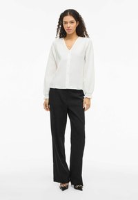 Hvid plisseret bluse med lange ærmer og V-hals, med knaplukning. Kombineret med sorte bukser med brede ben og spidse sorte sko.