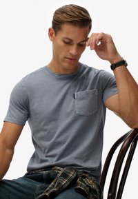 Grijze katoenen t-shirt met een ronde hals en korte mouwen, voorzien van een borstzak. G gedragen met donkere jeans en een geruit hemd om de taille geknoopt.