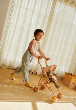 Plasticen kinderwagen in zacht roze met een gestreept stoffen zitje. Een kind in gestreepte tuinbroek duwt het, met een knuffeldier erin. Beige gordijnen op de achtergrond.