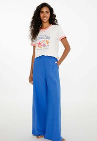 Bílá tričko s grafikou moře, růžovým límečkem a krátkými rukávy; spárováno s volnými, jasně modrými širokými kalhotami s visačkou.