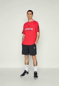 Vyras stovi vilkėdamas raudoną „Adidas“ sportinę marškinių, juodas „Adidas“ šortus, baltas „Adidas“ kojines ir juodas „Adidas“ sportines batų avalynes, fono paprastas.