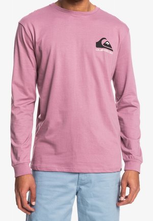 Homme portant un t-shirt à manches longues rose Quiksilver avec un logo noir et blanc sur la poitrine gauche et un pantalon bleu clair.