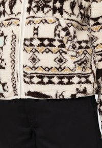 Polarowa kurtka w kremowej bazie, z geometrycznymi wzorami w kolorze czarnym i żółtym. Zamek błyskawiczny, boczne kieszenie i teksturowane wykończenie.