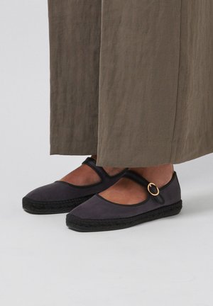 Ballerines Mary Jane noires avec une boucle dorée, portées avec un pantalon large olive, debout sur un sol gris clair.