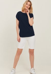 Mørkeblå kortærmet top i blødt stof, parret med hvide denimshorts med rå kanter. Model iført hvide sneakers. Enkel design.