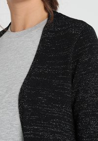 Cardigan nero lavorato a maglia con un motivo testurizzato, arricchito da fili argentati, indossato sopra una maglietta grigia chiara.