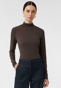 Brauner Langarm-Turtleneck-Shirt aus weichem Stoff, kombiniert mit dunkelblauen, maßgeschneiderten Hosen mit einer Fronttasche.