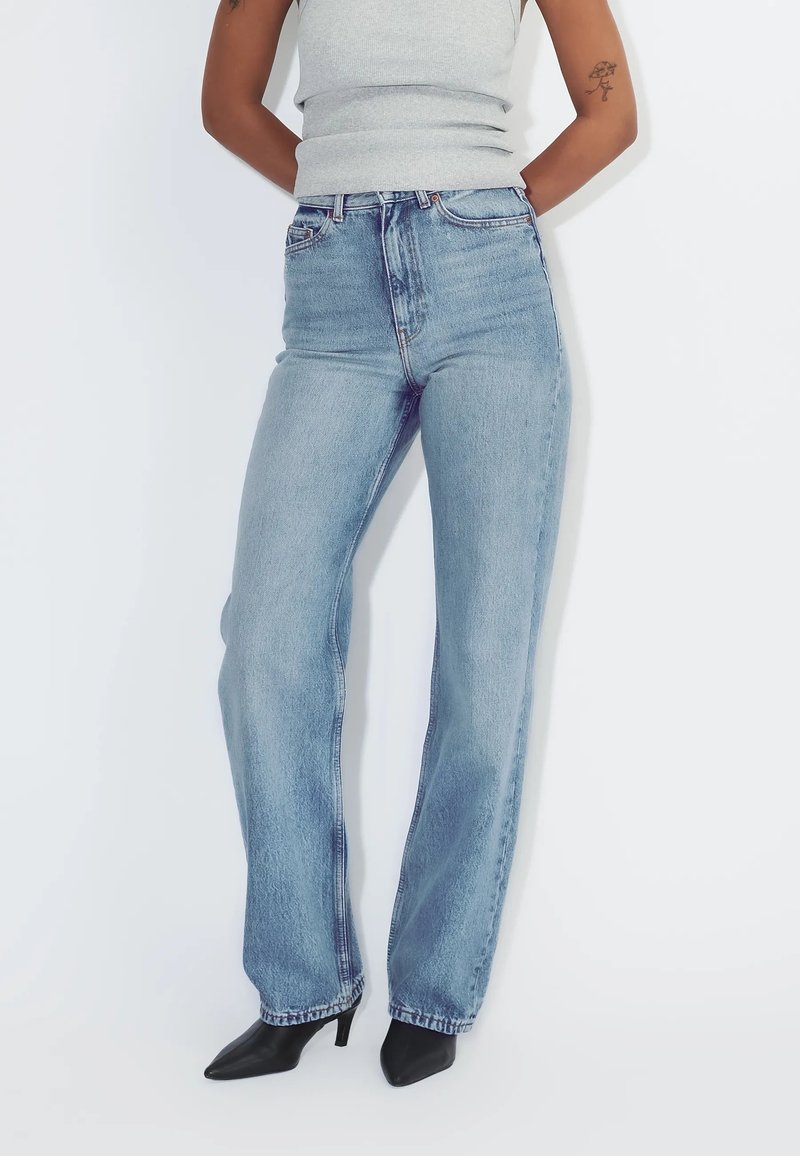 Person iført lyseblå højtaljede, lige jeans, grå ærmeløs top, sorte spidse hæle, stående med hænderne bag ryggen.