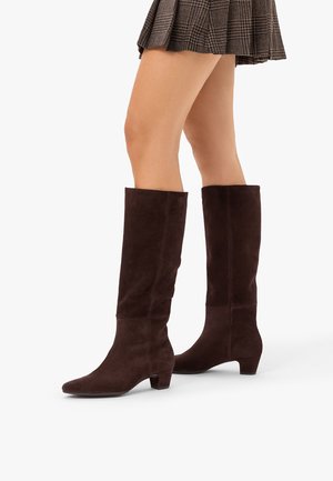 Braune kniehohe Wildlederstiefel, getragen mit einem braunen karierten Faltenrock an einer Person, die vor einem weißen Hintergrund steht.