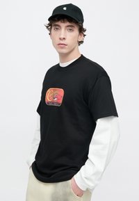 Camiseta negra de manga corta con un gráfico de estilo vintage en el frente, sobre una camisa blanca de manga larga, combinada con pantalones de color claro.