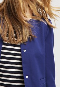 Blauwe cardigan met knoopsluiting en twee voorzakken, gedragen over een zwart-wit gestreept topje met korte mouwen. Gladde stoftextuur.