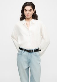 Νέ woman με σγουρά μαλλιά, φοράει ένα λευκό πουκάμισο με κουμπιά, κεντημένο μέσα σε ανοιχτό μπλε τζιν με μαύρη ζώνη.