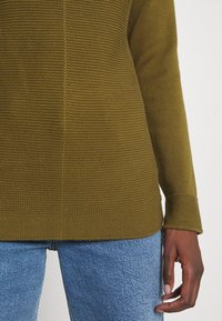 Maglione a maglia verde oliva con una finitura testurizzata, maniche lunghe e orlo dritto. Indossato con jeans in denim blu chiaro.