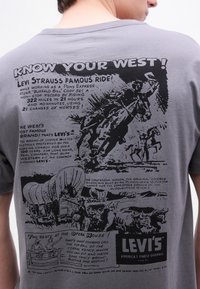 Πίσω μέρος γκρι πουκάμισου με μαύρες εικονογραφήσεις vintage στυλ που απεικονίζουν καουμπόηδες, άλογα, έναν καλυμμένο  άμαξα, και κείμενο σχετικά με τον Levi Strauss και την ιστορία της Δύσης.