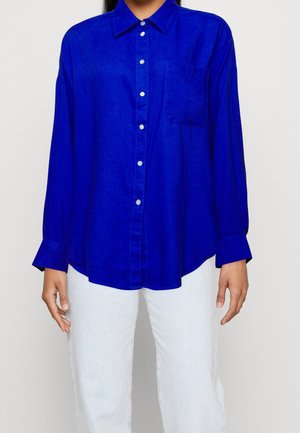 Kobieta w jasnoniebieskiej koszuli z guzikami, kieszenią na piersi i długimi rękawami, w połączeniu z jasnoniebieskimi dżinsami, na gładkim tle.