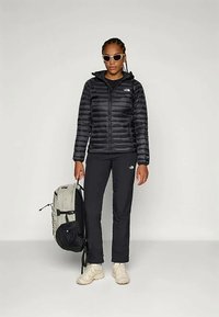 Μαύρο μπουφάν με μόνωση, οριζόντιες ραφές, μπροστινό φερμουάρ και κουκούλα; σε συνδυασμό με μαύρο παντελόνι, λευκά sneakers και σακίδιο σε μπεζ/μαύρο χρώμα.