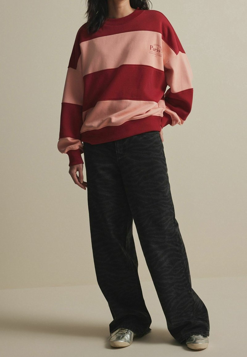 Personne portant un sweat-shirt rayé rouge et rose oversize, un pantalon large noir avec un motif discret et des baskets argentées, debout devant un fond uni.