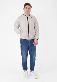 Giacca a vento grigio chiaro con cappuccio e zip frontale, dettagli gialli e blu. Abbinata a jeans in denim blu e sneakers bianche.