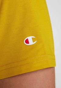 Yellow cotton t-shirt fabric with a smooth texture, featuring a white and red Champion logo on the left side and yellow stitching.
