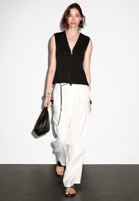Donna che cammina al chiuso indossando un gilet nero senza maniche, pantaloni bianchi larghi, sandali neri, e tiene una borsa nera nella mano destra.