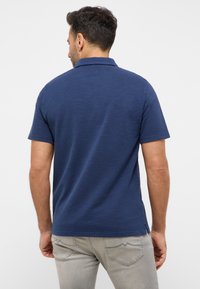 Jūriniai polo marškiniai su trumpomis rankovėmis, tekstūruota medžiaga ir klasikinis apykaklė; šonuose plyšiai tamprėje, derinami su šviesiai pilkais džinsais.