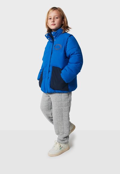 Blå puffjacka med en marinblå ficka, hög krage och grå kviltade byxor. Kombinerad med vita sneakers som har subtila varumärkesdetaljer.