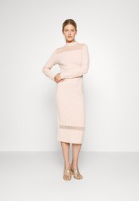 Vestido rosa claro feito de tecido suave, apresentando mangas longas e um detalhe de renda recortada no peito e na bainha. Comprimento midi.