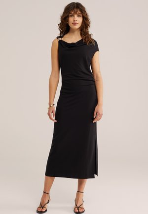 Femme aux cheveux bouclés portant une robe midi noire sans manches avec une fente latérale, un bracelet doré et des talons noirs à brides sur un fond clair.