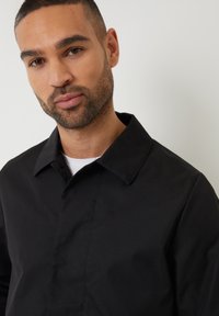 Černá bunda s límečkem a hladkou texturou, s knoflíkovým zapínáním a minimalistickým designem, nošena přes bílou košili.