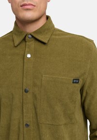 Camicia in corduroy verde oliva con colletto a punta, apertura con bottoni e taschino sul petto. Presenta bottoni argentati e un'etichetta del marchio tessuta.