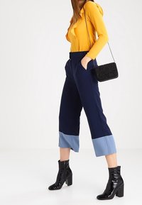 Tmavě modré zkrácené kalhoty se světle modrými manžetami, doplněné hořčicovým topem s dlouhým rukávem. Doplňky zahrnují texturovanou černou kabelku a lesklé černé boty.