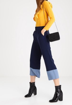 Tmavě modré zkrácené kalhoty se světle modrými manžetami, doplněné hořčicovým topem s dlouhým rukávem. Doplňky zahrnují texturovanou černou kabelku a lesklé černé boty.