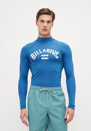 Hosszú ujjú kék búvárruházat, amelyen fehér "Billabong" logó található a mellkasán, rugalmas, vízálló anyagból készült.